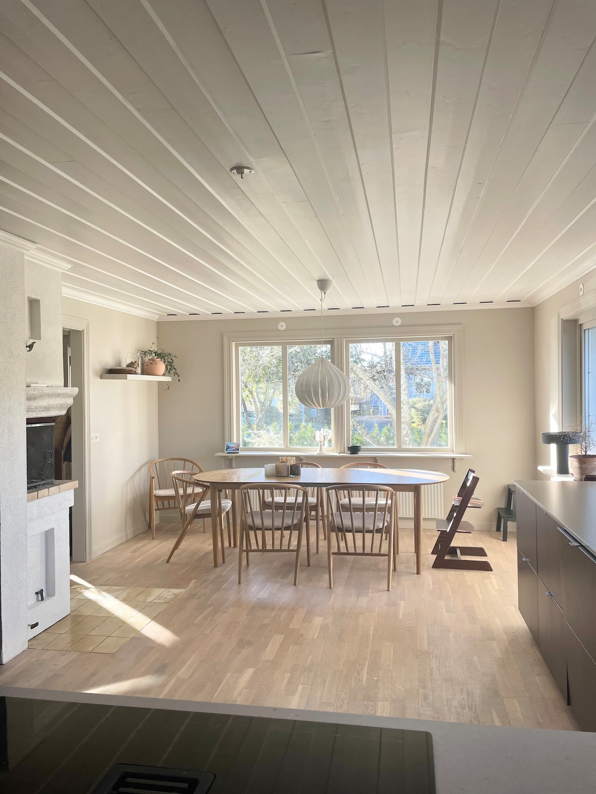 A well-lit dining area features a round table surrounded by several wooden chairs. Large windows provide natural light and views of the greenery outside. A pendant light hangs overhead, and minimalistic decor adds a contemporary touch to the space.