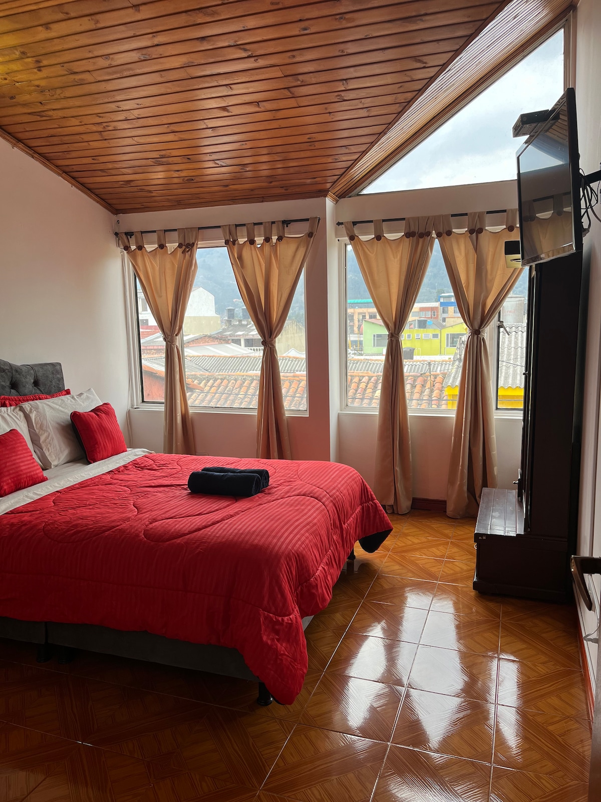 A well-lit bedroom features large windows, dressed in light curtains, allowing natural light to brighten the space. A spacious bed, covered with a red quilt, is paired with red accent pillows. A television is mounted on the wall opposite the bed, contributing to a functional layout.