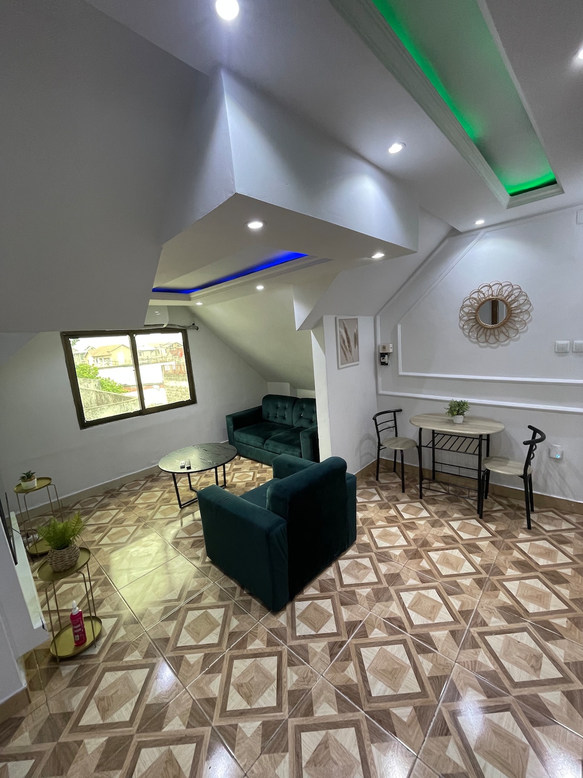 A well-lit living area is furnished with a green sofa and a glass coffee table. A round dining table with metal chairs is positioned near a window, allowing natural light to fill the space. Decorative elements include a wall mirror and indoor plants.