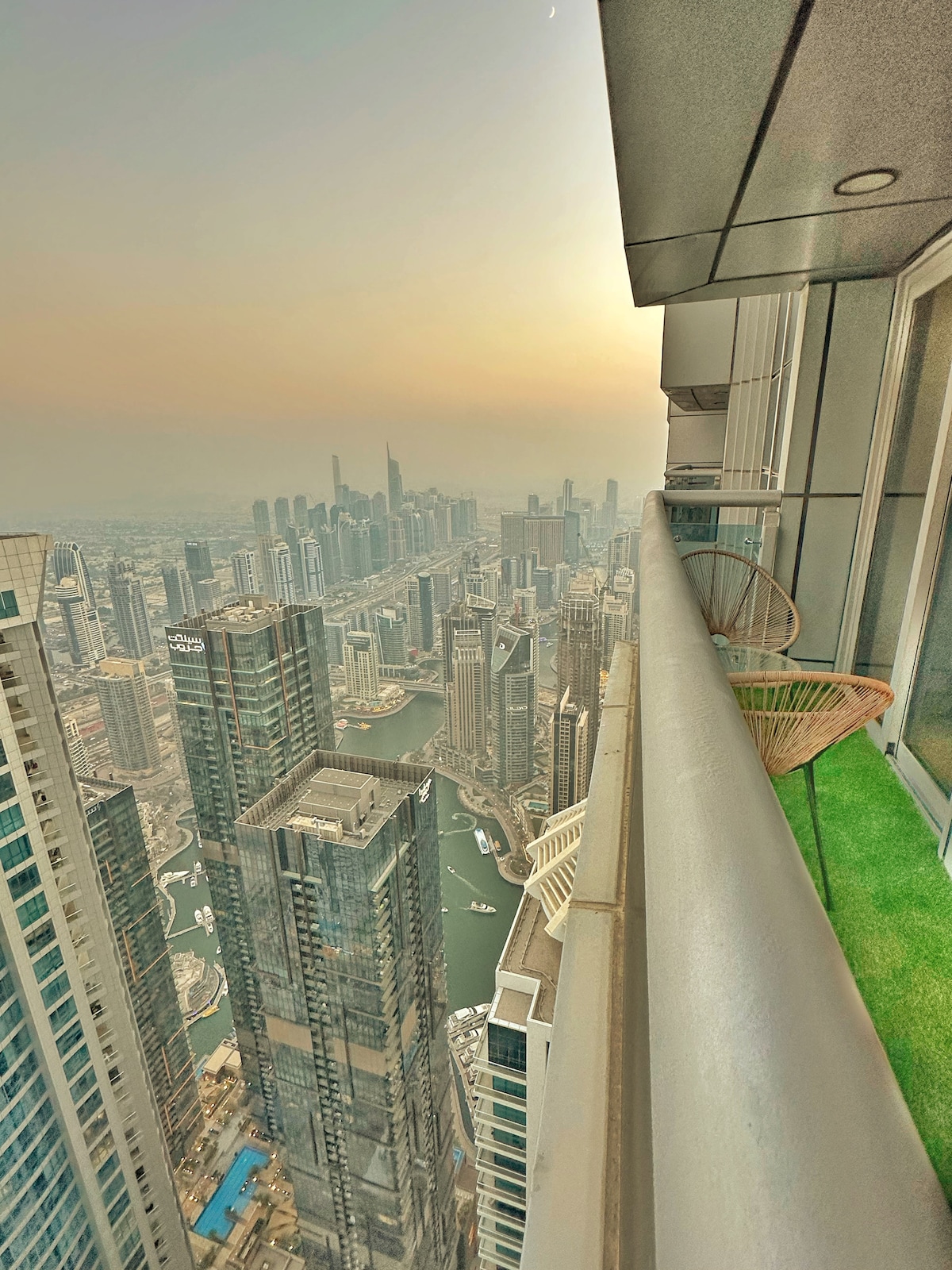 The balcony offers a panoramic view of the city skyline, showcasing a mix of modern skyscrapers and the winding waterfront. Two chairs are positioned on the green carpet, inviting relaxation while overlooking the bustling landscape below, illuminated by the setting sun.