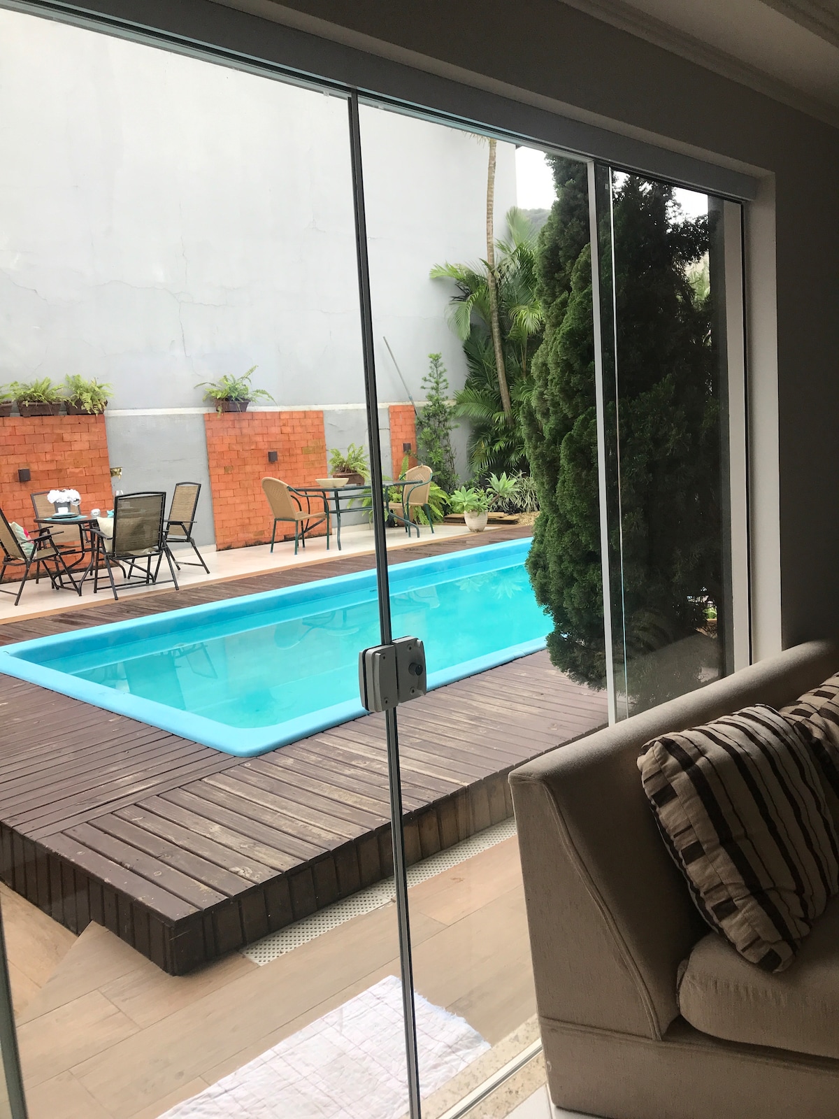 A clear view of a pool area is presented through large glass doors. The sparkling blue water is surrounded by a wooden deck and potted plants. A seating area with chairs and tables is visible, creating an inviting outdoor space.