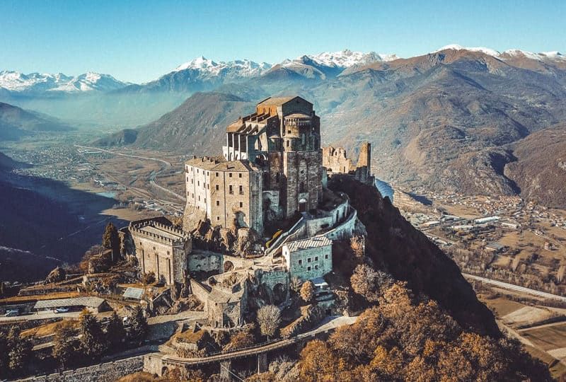 A historic castle is perched on a mountain, surrounded by expansive valleys and distant snow-capped peaks. The structure showcases a mix of stone and wooden elements, with winding pathways leading through the landscape. The clear sky enhances the scenic beauty of the alpine surroundings.