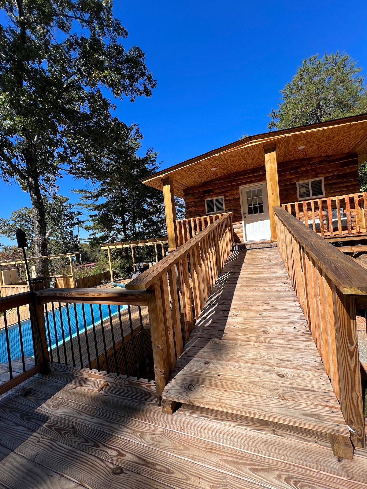 A wooden deck leads to a charming cabin showcasing a welcoming entrance. The surrounding environment features tall trees, casting shadows on the pathway. A sparkling swimming pool is visible to the left, adding to the outdoor appeal.