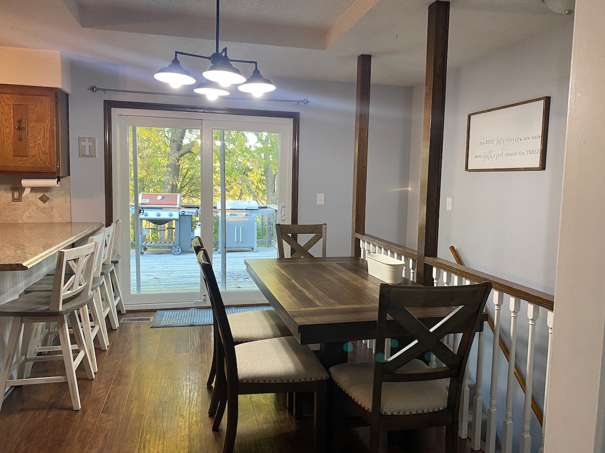 A dining area features a wooden table with seating for six, complemented by soft-toned fabric chairs. Light from a modern chandelier illuminates the space, while large glass doors provide access to the outdoor deck, enhancing the connection to nature.