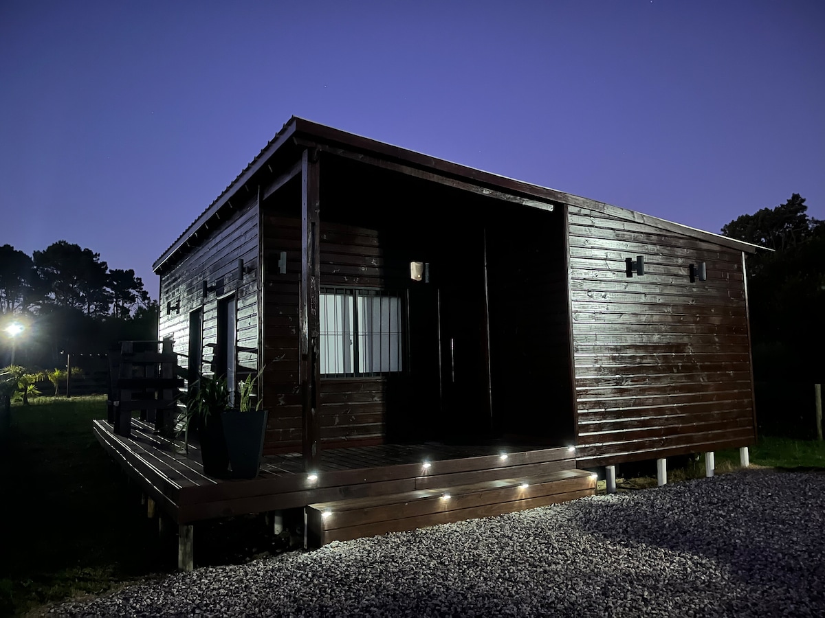 The exterior of a wooden cabin is illuminated by soft outdoor lighting, highlighting its robust structure. A gravel pathway leads to a covered deck, offering seating options. Surrounding greenery adds a natural backdrop, while the night sky is visible above.