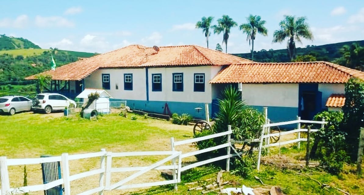 A traditional country house with a red-tiled roof and whitewashed walls is surrounded by a spacious lawn. Palm trees and lush greenery are visible in the background, contributing to a serene outdoor environment. Several vehicles are parked nearby, adding to the rustic charm.