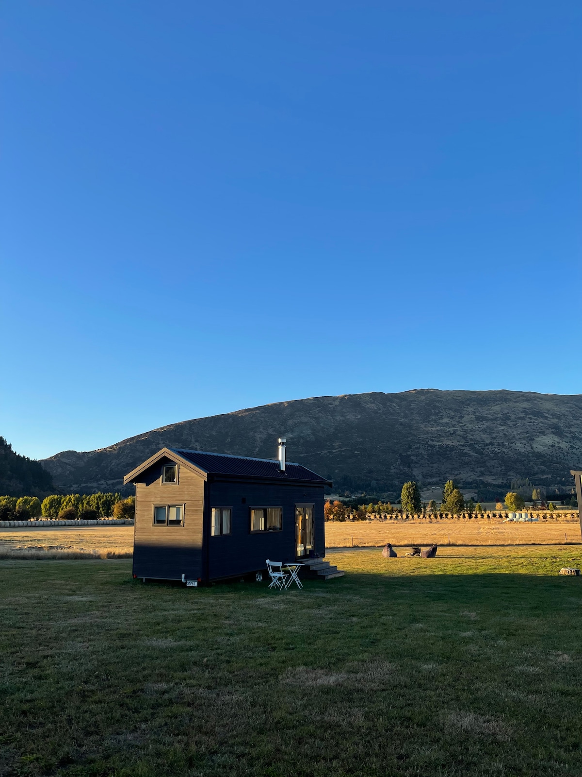 The tiny house is set against a backdrop of rolling hills under a clear blue sky. The structure features a dark exterior, a chimney, and a small porch with two chairs, surrounded by a grassy area that offers a peaceful retreat.