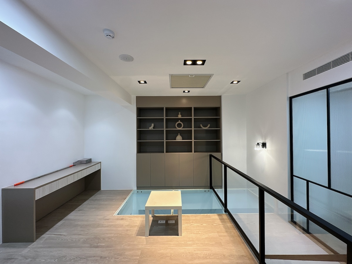 An open, airy space features a bright floor with light wood planks and modern furnishings. A low table is positioned over a glass-enclosed area, complemented by a sleek shelving unit with decorative items. Natural light enters through large windows, highlighting the minimalist design.