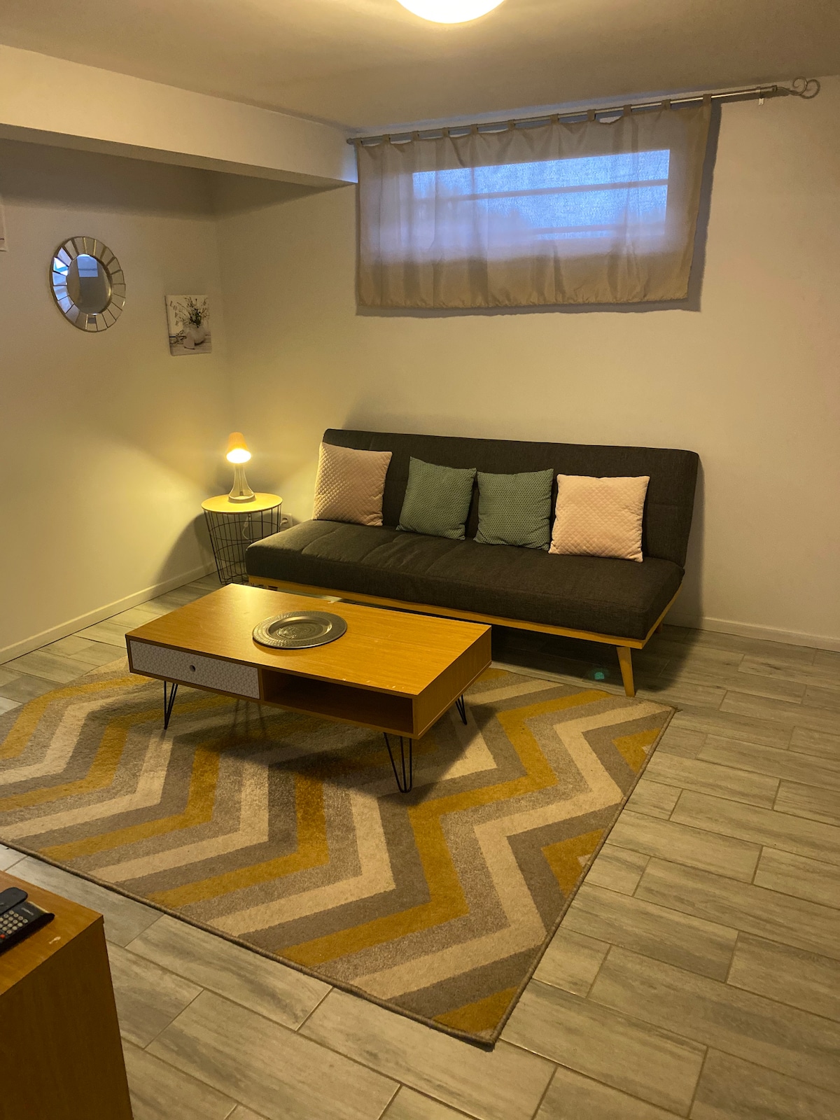 A cozy living area features a modern gray sofa adorned with decorative cushions. A wooden coffee table rests on a patterned rug in warm tones. A table lamp provides soft lighting, and a round mirror is mounted on the wall, reflecting the serene ambiance.