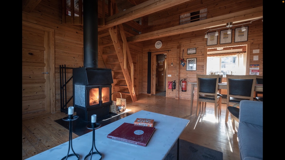 A spacious living area features wooden walls and flooring, highlighted by a cozy wood-burning stove. A dining table with chairs is placed near large windows, while a coffee table with decor items and candles enhances the atmosphere. A staircase leads to the upper level.
