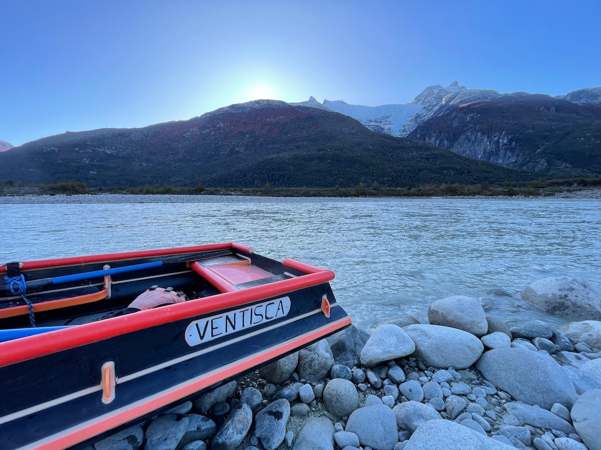 A boat named 'Ventisca' rests on rocky shores beside a river, with swirling water reflecting the surrounding landscape. Majestic mountains rise in the background, partially covered by snow, beneath a clear sky with the sun setting on the horizon.