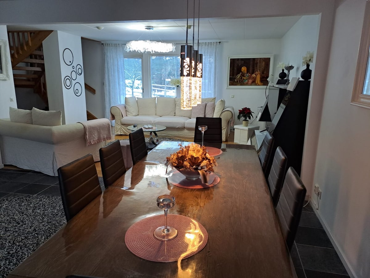 A dining table is set with an arrangement of decorative items, surrounded by dark chairs. A spacious living area is visible in the background, featuring a light-colored sofa and large windows that allow natural light to fill the space. Unique hanging light fixtures add visual interest.