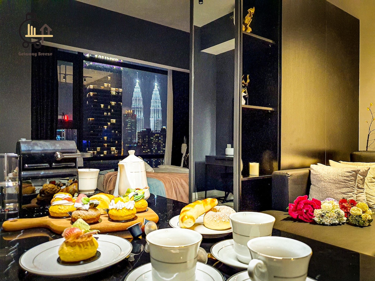 A dining table is set with white tea cups and an assortment of pastries, including colorful desserts. In the background, floor-to-ceiling windows provide a view of the iconic Petronas Twin Towers, enhancing the urban ambiance. A cozy bed and modern furnishings complement the scene.