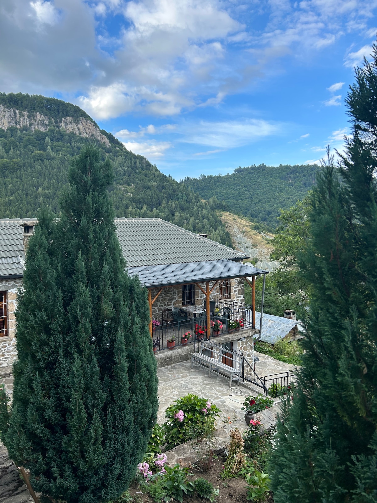A charming stone building is surrounded by lush greenery and tall evergreen trees. The structure features a gray roof and a welcoming porch adorned with colorful flowers. Rolling hills and a clear blue sky create a serene backdrop.