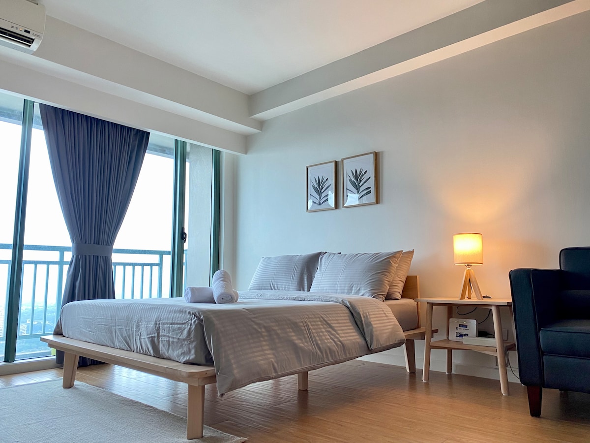 A spacious bedroom features a queen-sized bed with a premium memory foam mattress. Light-colored bedding contrasts with the light wood furniture. A lamp and a small side table are positioned beside the bed, while a two-seater sofa is placed in the corner. Large windows are partially covered by soft blackout curtains.