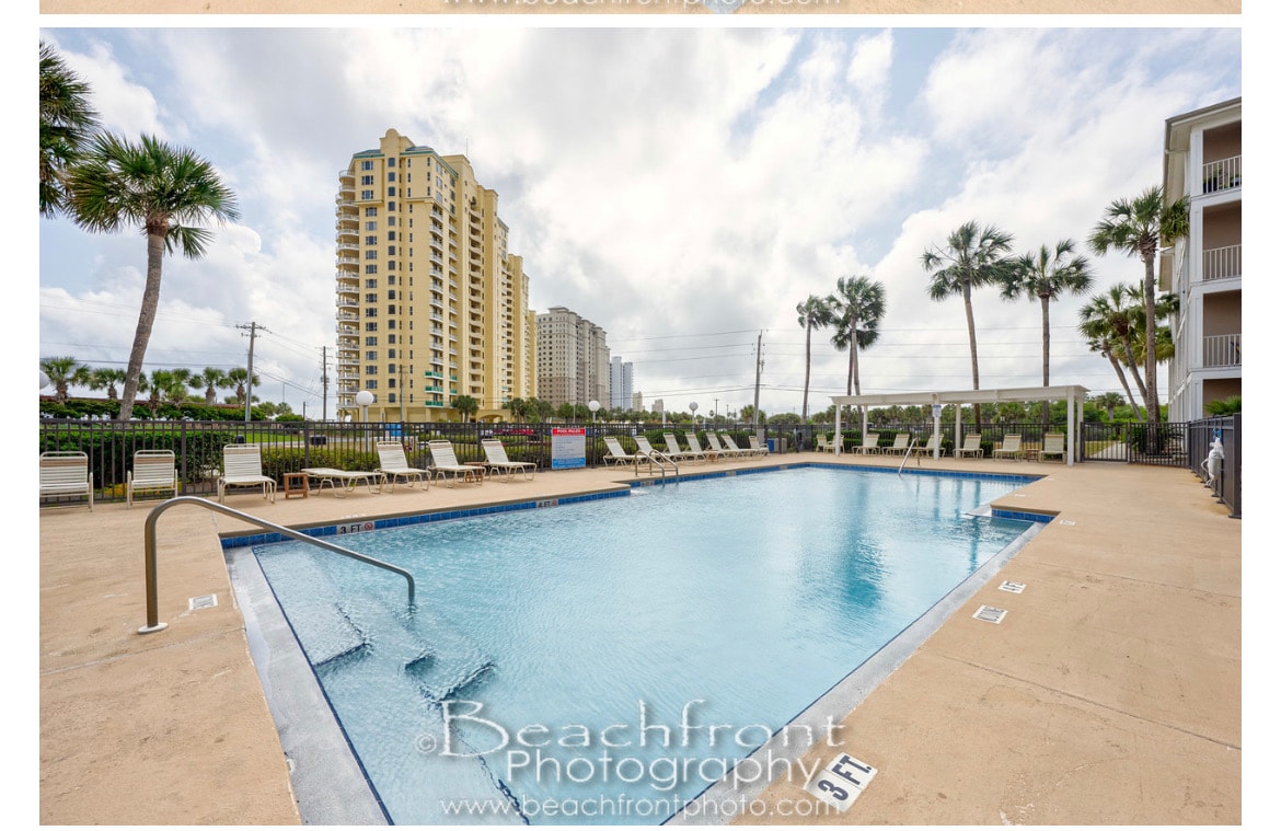 The outdoor swimming pool is outlined by a spacious deck featuring multiple lounge chairs. Palm trees provide a tropical touch, while tall buildings rise in the background against a cloudy sky. The pool area is securely fenced, indicating a well-maintained recreational space.