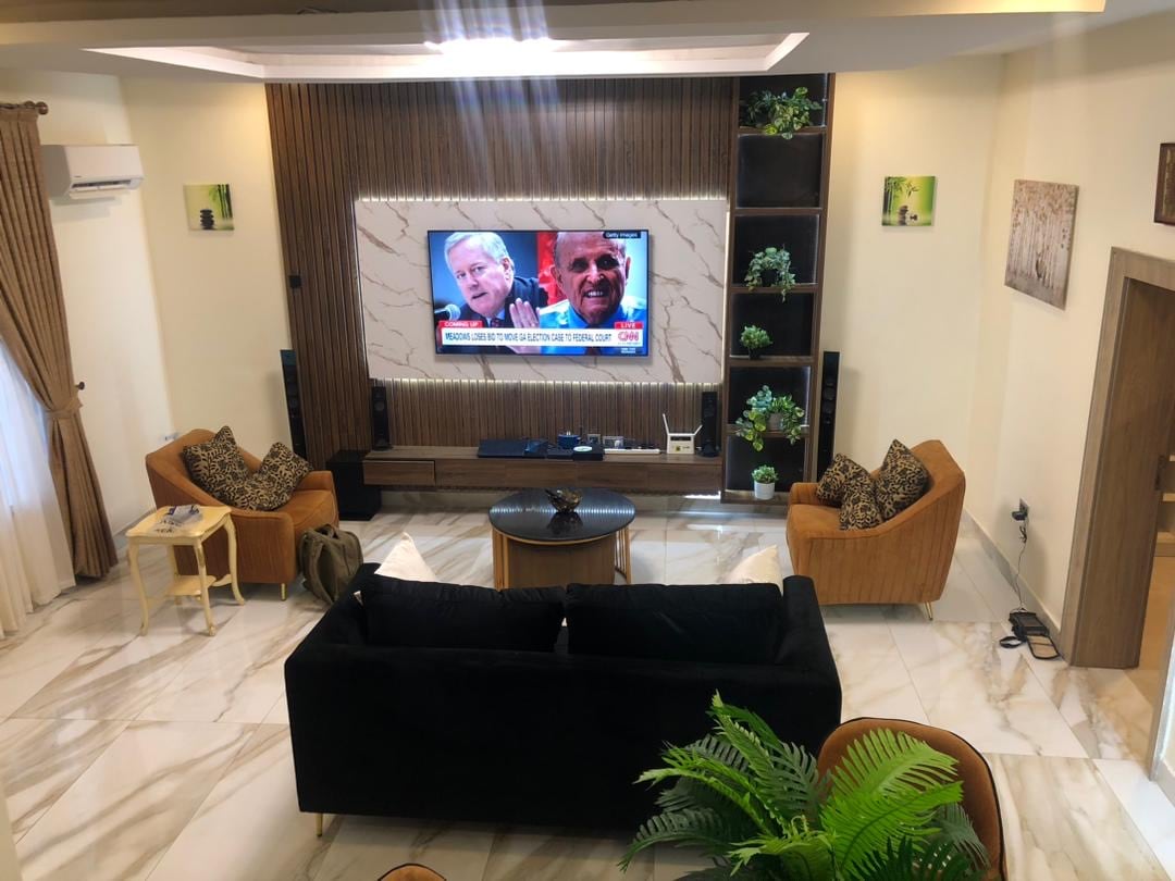 A spacious living area features a large flat-screen TV mounted on a decorative wall. Comfortable armchairs in warm tones are complemented by a round coffee table. Decorative plants and artwork enhance the ambiance, while large windows provide natural light.