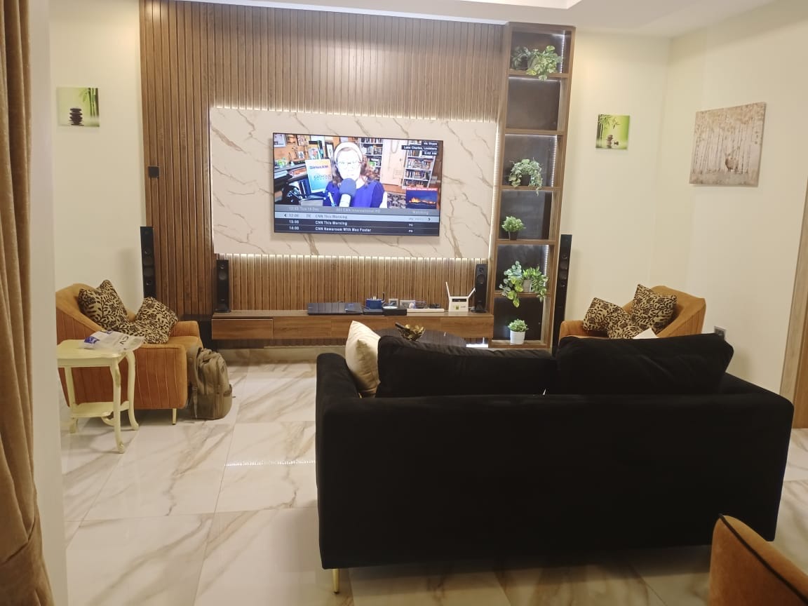 The living area features a modern design with a large flat-screen TV mounted on a marble accent wall. Comfortable seating is provided by a black sofa and two brown armchairs adorned with decorative cushions. Greenery accents are placed on shelves, adding a touch of nature.
