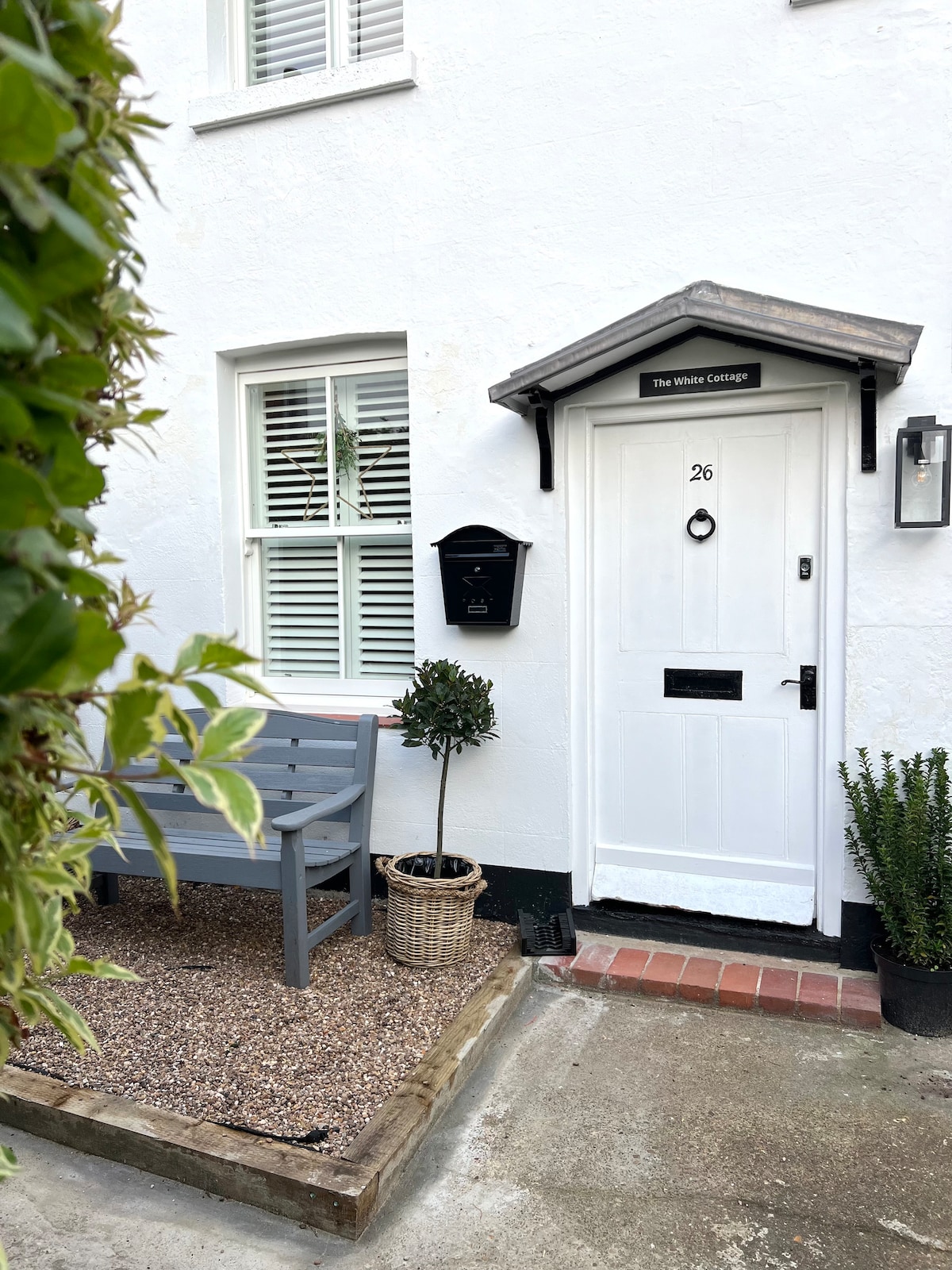 The entrance to The White Cottage is framed by a small gravel area, featuring a bench and potted plants. A white door with a prominent number 26 is accompanied by a mailbox. Wooden shutters enhance the welcoming appearance of the entry.