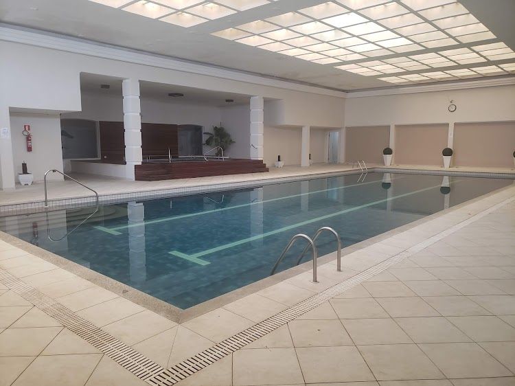An indoor swimming pool is surrounded by large windows, providing natural light and a serene environment. The pool features a tiled border with steps leading into the water. Comfortable seating areas are placed nearby, enhancing the relaxation potential within the space.