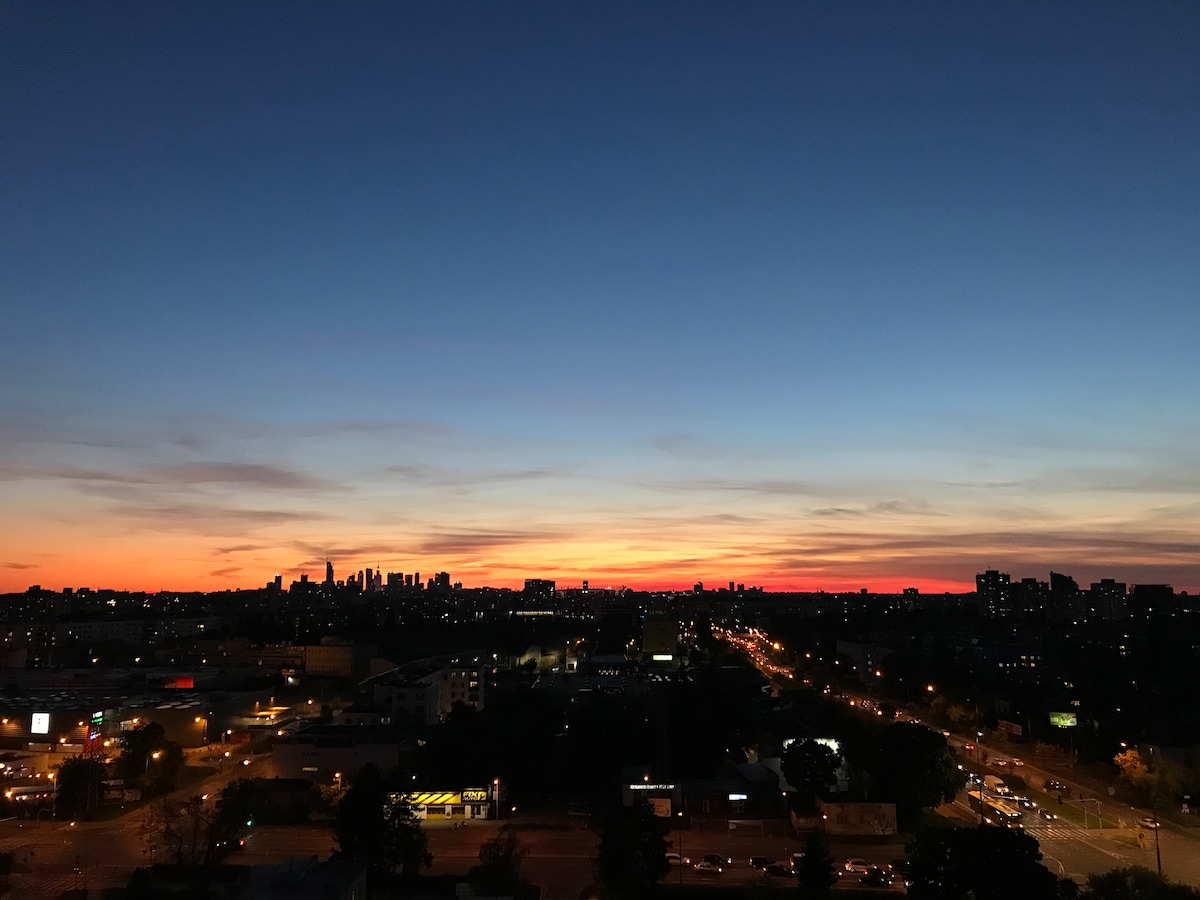 A stunning urban skyline is displayed at dusk, with a rich gradient of orange, purple, and blue hues painting the horizon. The outline of city buildings is visible against the vibrant sunset, while city streets below are illuminated with soft lights.