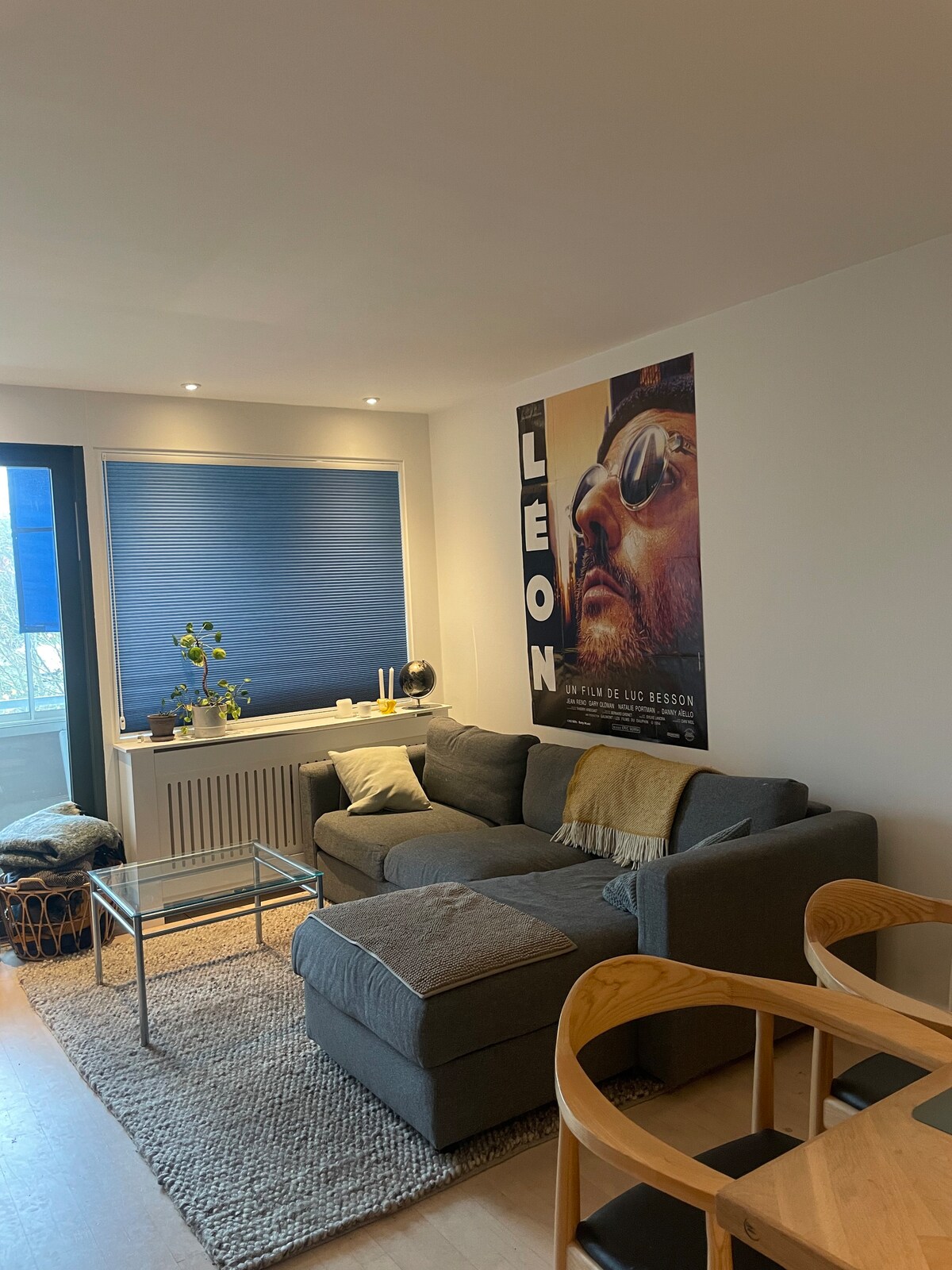 A modern living room features a spacious gray sectional sofa adorned with a beige throw blanket. A glass coffee table sits on a textured area rug, while a large window with blue shades provides natural light. Decorative plants and a stylish wall decoration add character.