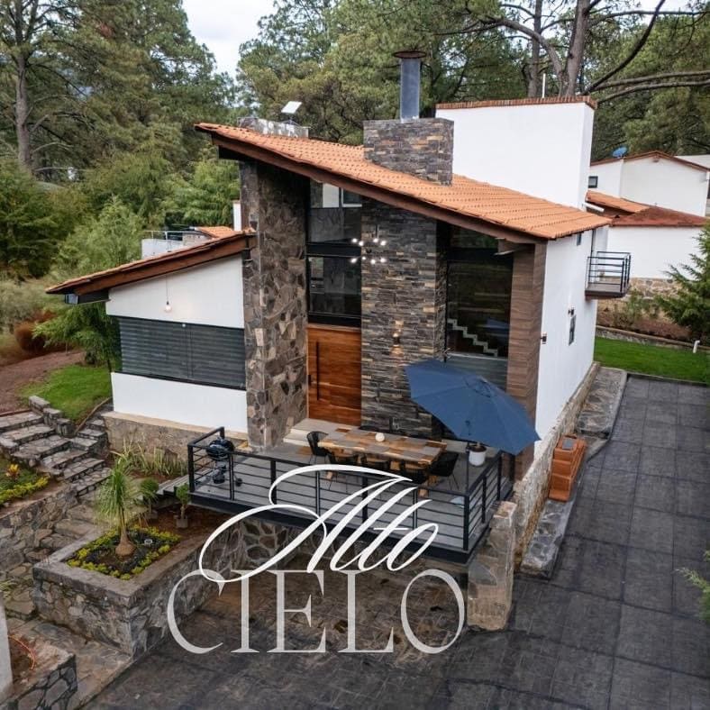 The exterior of Cabaña Alto Cielo features a blend of stone and modern architectural elements. A welcoming porch is adorned with an umbrella and overlooks a landscaped area. Natural surroundings are visible, contributing to an inviting atmosphere for relaxation.