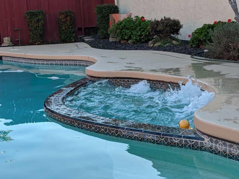 A private hot tub is integrated into the pool area, featuring bubbling water that suggests a relaxing atmosphere. The surrounding concrete deck is complemented by neatly trimmed greenery and vibrant flowers, enhancing the outdoor space's appeal.