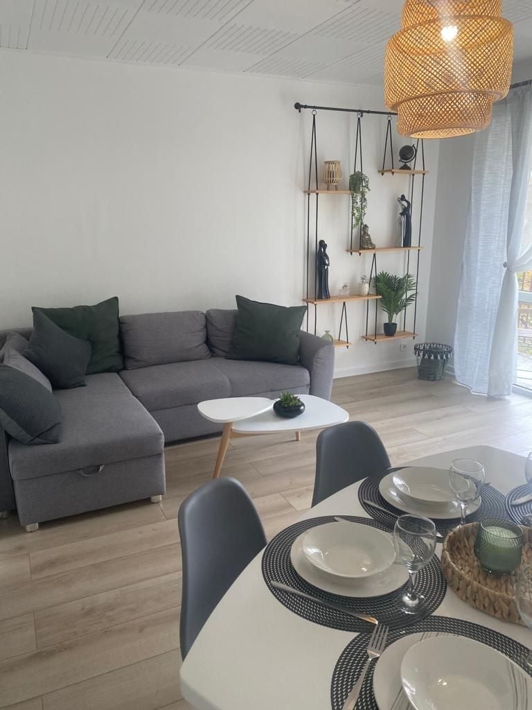 A gray sectional sofa is positioned in a sunlit living area, complemented by decorative pillows. A small white table sits in front of the sofa. A dining table, set with plates and glassware, features a woven centerpiece. A shelf with decorative items and plants is visible in the background.