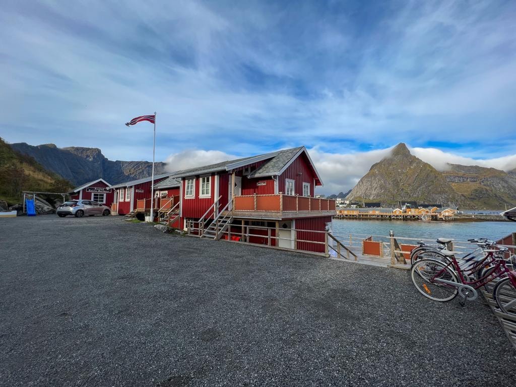 A charming red building is positioned by the water, with a Norwegian flag flying above. Surrounding mountains and a blue sky complete the scenic view. Outdoor seating and bicycles are available nearby, contributing to the welcoming atmosphere of the location.