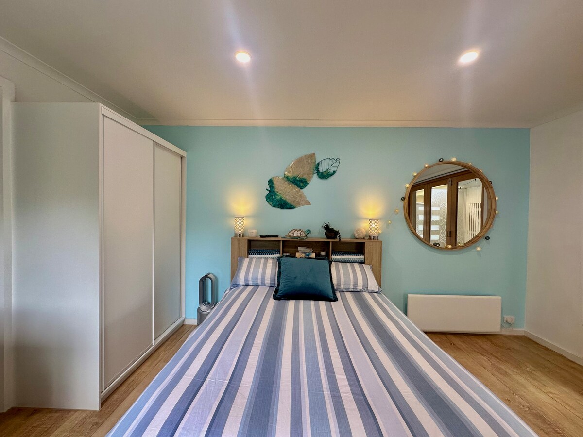 A cozy bedroom features a double bed with a striped blue and white duvet cover, accented by a solid blue cushion. A round mirror adorns the wall alongside decorative wall art, with soft lighting provided by two bedside lamps. A wardrobe is visible to the left.