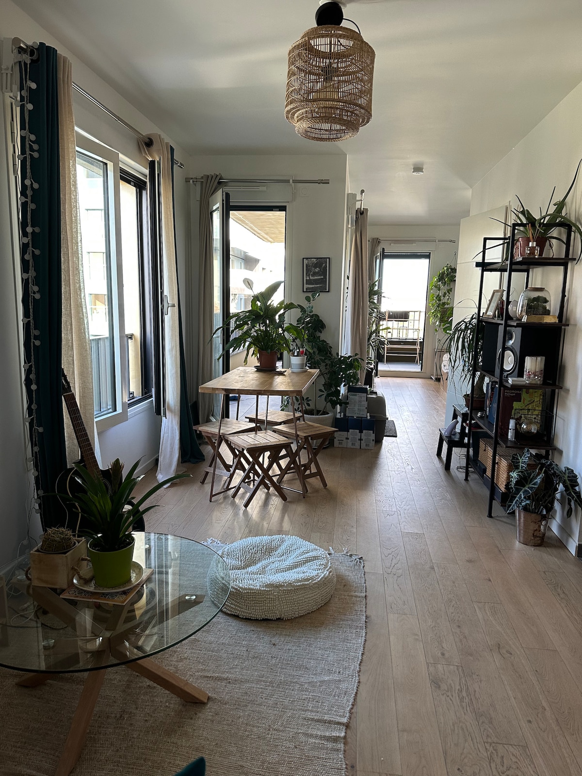 A bright living space features large windows with natural light streaming in. A round glass coffee table sits on a textured rug, accompanied by a comfortable pet bed. A wooden dining table with two chairs is positioned alongside potted plants, enhancing the welcoming atmosphere.