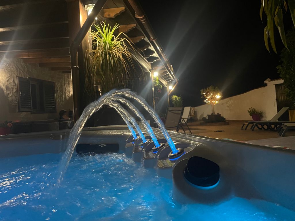 The jacuzzi is illuminated by blue lights, highlighting the flowing water. Surrounding the jacuzzi, lounge chairs are positioned on a tiled patio, and soft outdoor lighting creates a serene atmosphere. The structure overhead features wooden beams and hanging plants, enhancing the relaxing setting.