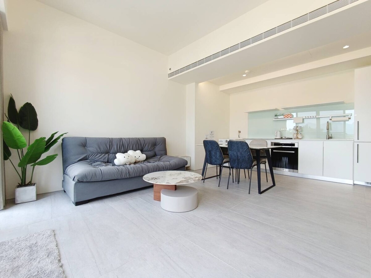 A modern living area is presented with a grey sofa arranged alongside a potted plant. A circular coffee table features a marble surface, complemented by a dining table with four dark chairs. The space is illuminated by natural light from nearby windows.
