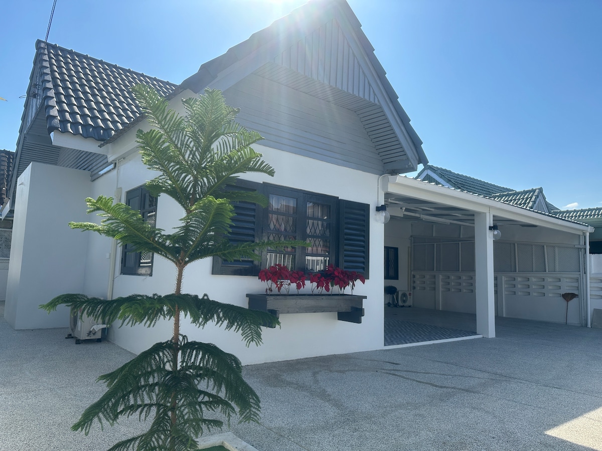 The exterior of a cozy house is showcased, featuring a peaked roof with gray tiles and white walls. A decorative planter with tropical plants is positioned in front, and a spacious driveway is visible, inviting guests to enter the welcoming space.