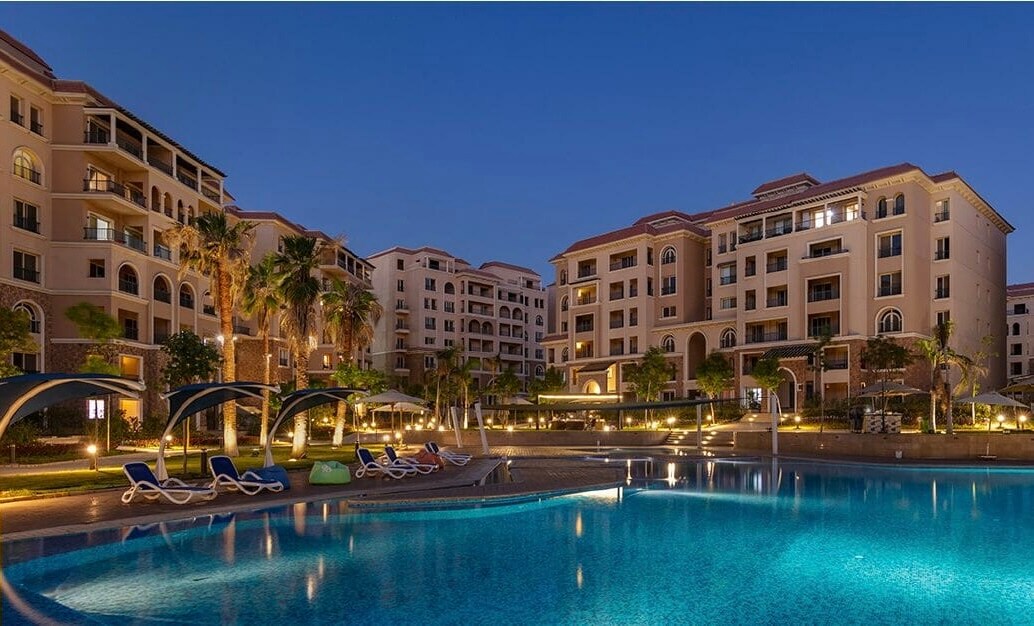 A well-lit pool area is surrounded by elegant buildings, showcasing a blend of modern architecture and greenery. Loungers are positioned near the water, while palm trees and subtropical plants provide natural accents. The scene is enhanced by soft lighting that reflects off the calm surface of the pool.