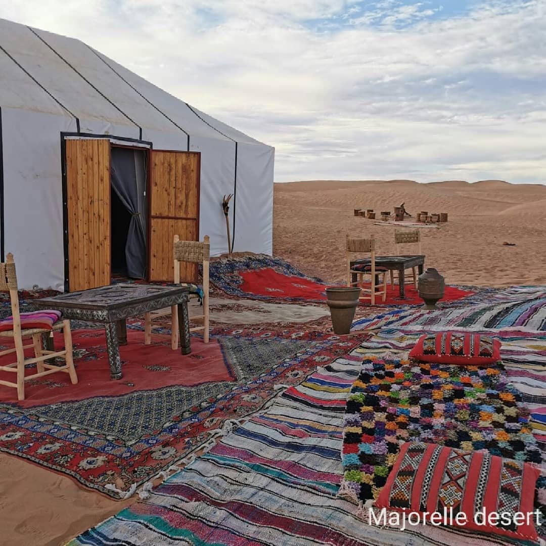 An inviting desert tent is accompanied by woven rugs and traditional floor seating. Several wooden chairs and a low table are positioned outside, set against the backdrop of rolling sand dunes. The sky displays soft clouds, enhancing the serene atmosphere.