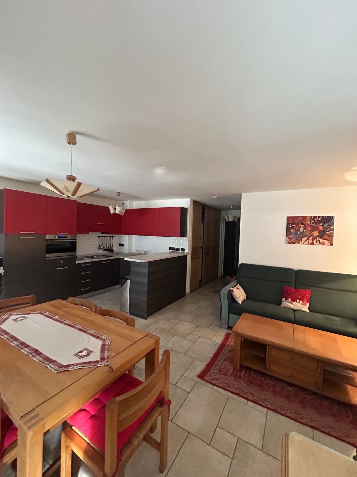 An open kitchen featuring red and gray cabinetry is visible, equipped with modern appliances. A dining table with wooden chairs and a comfortable seating area is present. Warm lighting enhances the space's ambiance, while a wall art piece adds a decorative touch.