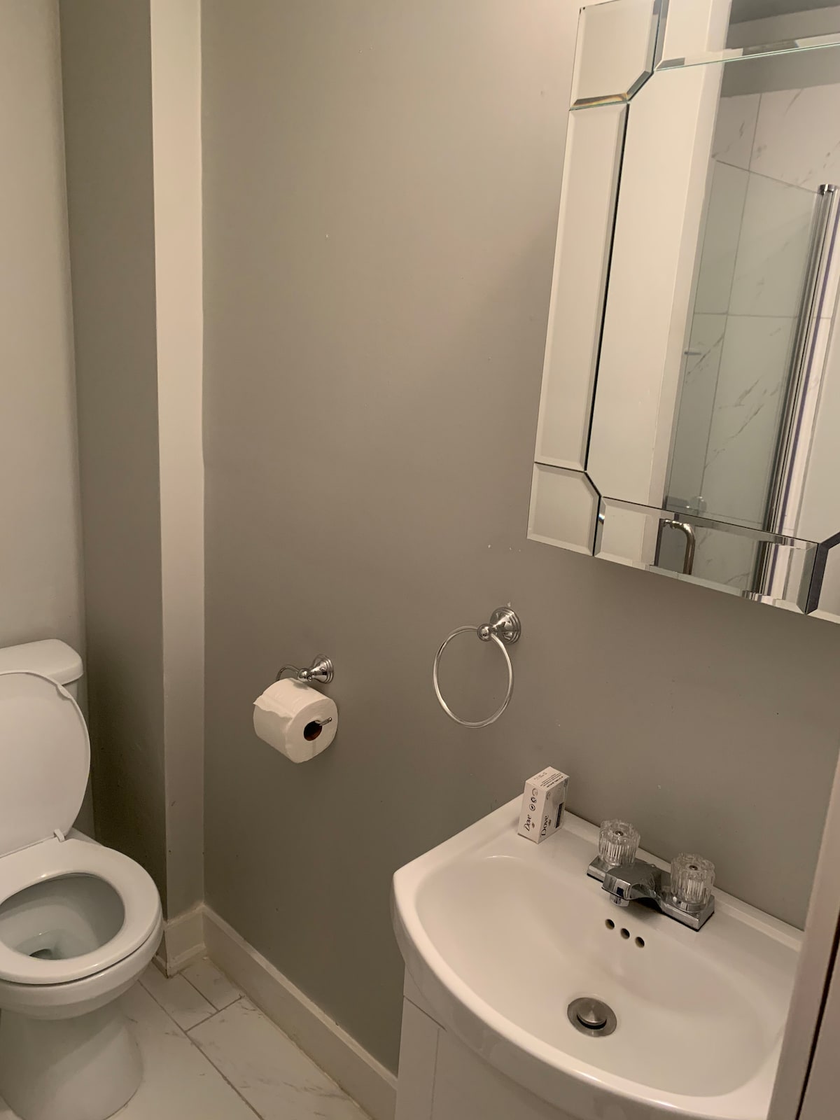 The compact bathroom features a modern toilet and a small sink accompanied by a stylish mirror. A towel ring and a roll of toilet paper are visible, highlighting the essential amenities for convenience. Light-colored tiles enhance the clean and simple design.