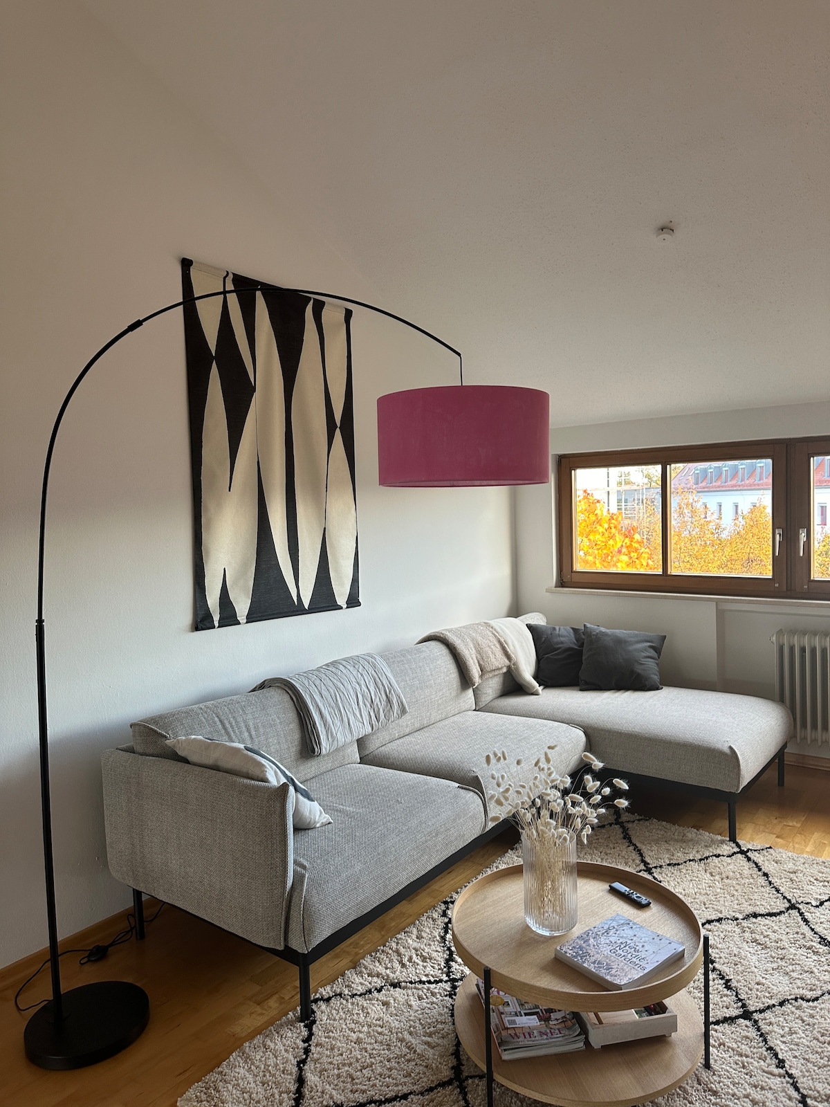 A cozy living area features a modern sectional sofa in neutral tones, complemented by a pink floor lamp. Natural light enters through the large windows, which overlook a colorful autumn scene. A round coffee table, adorned with a vase and magazines, completes the setting.