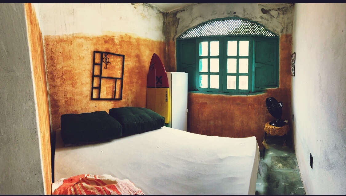 A room is presented with an orange and beige textured wall. A spacious bed rests against one wall, complemented by two dark green pillows. A wooden cabinet and a small fan are visible, next to a window with distinctive green shutters, providing natural light.