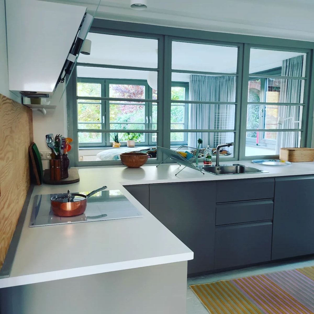 A modern kitchen space is showcased, featuring sleek gray cabinets and a white countertop. Cooking utensils are neatly arranged, with a pot on the stovetop. Large windows provide a view into the garden, allowing natural light to fill the area.