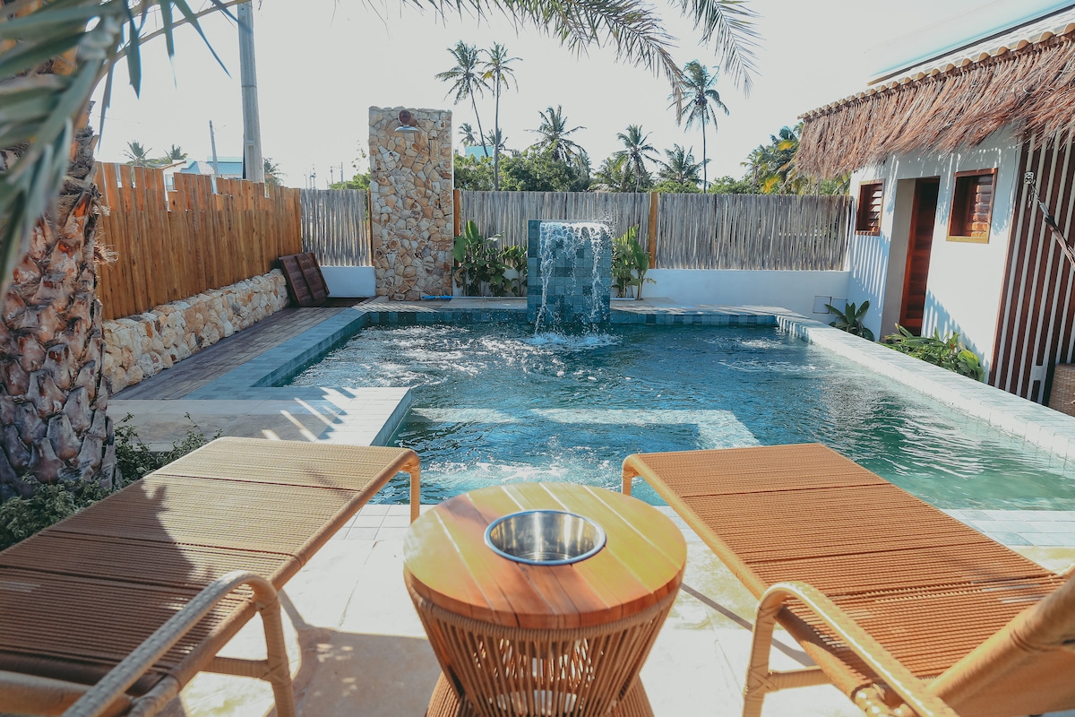 A serene outdoor area features a shimmering pool with a gentle waterfall, surrounded by lush greenery and wooden fencing. Two lounge chairs are positioned beside a wooden table, offering a relaxing spot to enjoy the tranquility of the space.