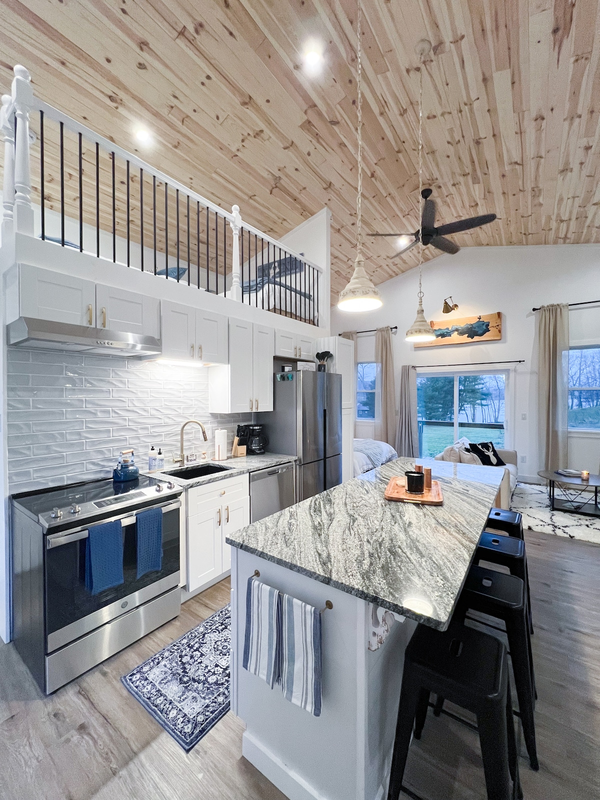 A modern kitchen is displayed, featuring stainless steel appliances and a granite island with seating. Natural light is provided by large windows, illuminating the space. The high wooden ceiling enhances the open feel, while decorative elements add character to the area.