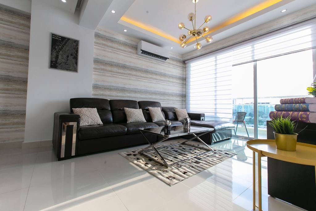 A modern living area showcases a sleek black sofa adorned with decorative cushions. A stylish coffee table is positioned on a textured rug, while a large window allows natural light to illuminate the space. Minimalist decor enhances the contemporary design.