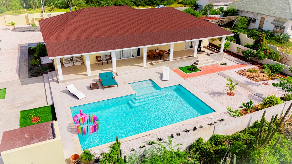 An aerial view of the villa reveals a large turquoise pool with a shallow area and vibrant pool floats. The spacious patio features lounge chairs and a dining area. Surrounding gardens with tropical plants and decorative stones enhance the outdoor space, while the red-tiled roof complements the overall aesthetic.