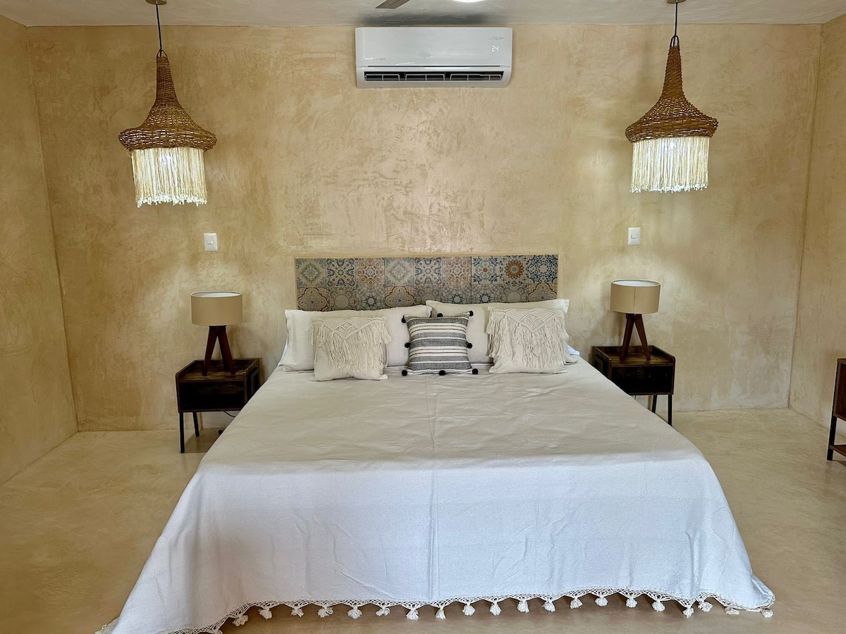 A spacious bed with a white bedspread and illuminated by two bedside lamps is centered in the room. Woven pendant lights hang from the ceiling, creating ambient lighting against the textured beige walls.