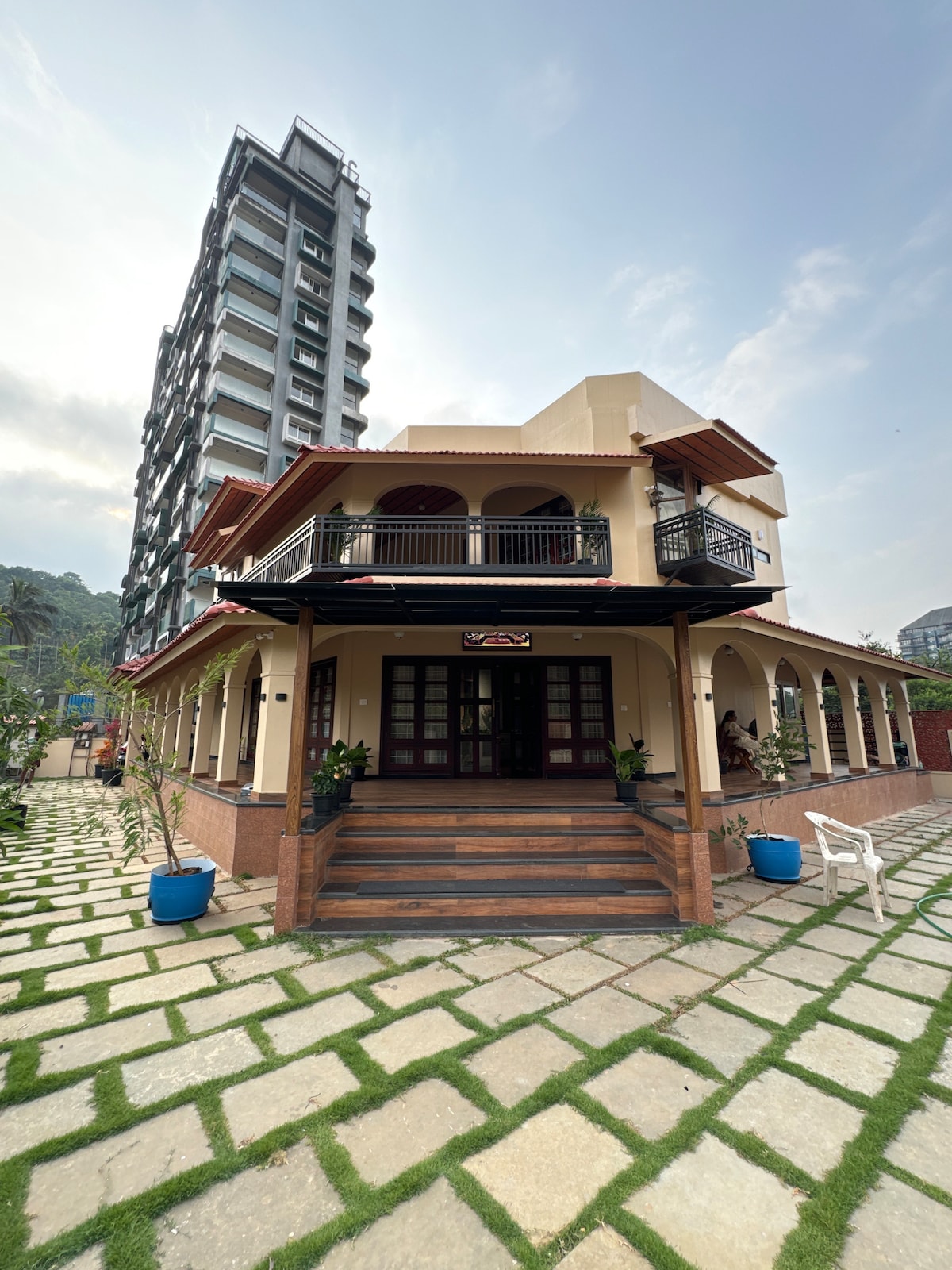 A charming two-story structure is featured, surrounded by a well-maintained garden with geometric stone pathways. A modern building looms in the background. The entrance is marked by wide stairs and a balcony adorned with potted plants, showcasing a blend of contemporary and traditional architecture.