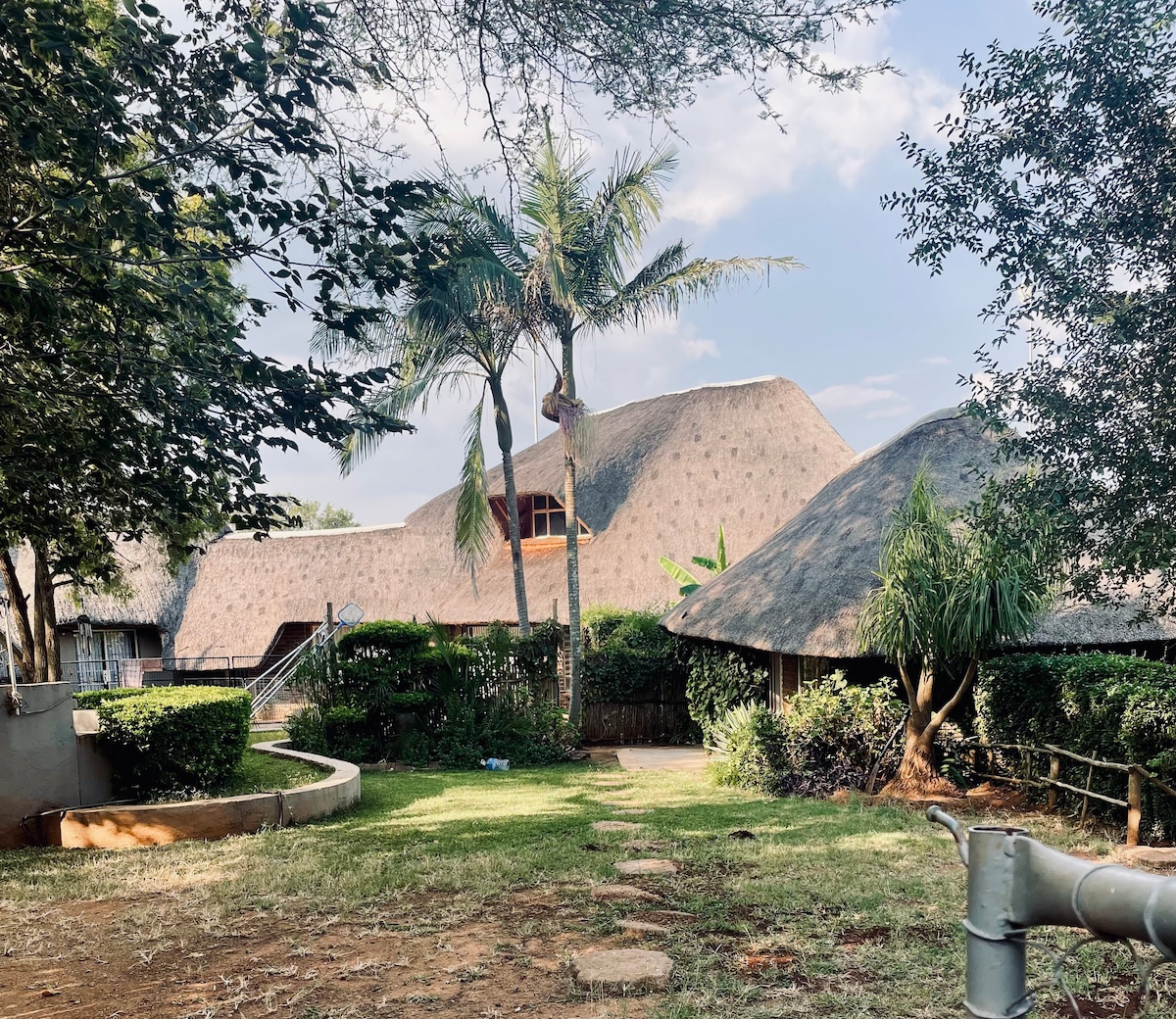 A rustic farmhouse is set within a lush garden, featuring thatched roofs and landscaped greenery. Pathways lead through the area, surrounded by palm trees and shrubs, creating a peaceful and inviting outdoor environment.