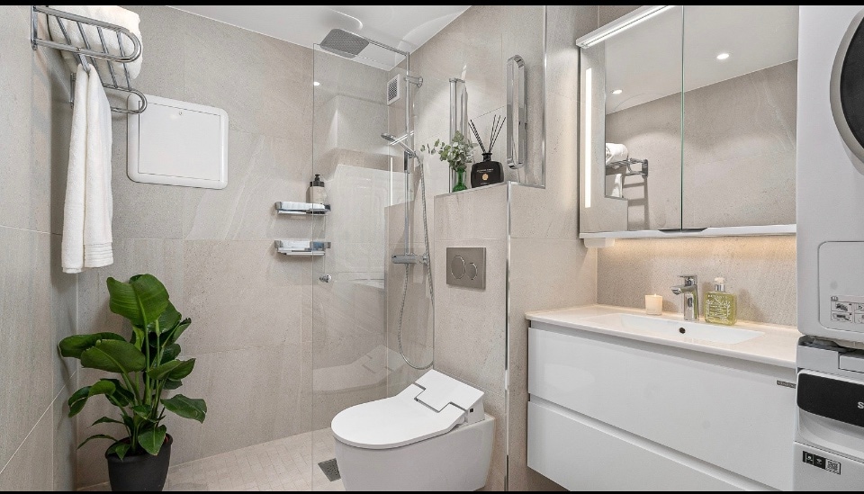 A modern bathroom features a sleek design with light-colored tiles and an illuminated mirror. A shower area is equipped with glass panels, and storage shelves hold various toiletries. A potted plant adds a touch of greenery to the space, which includes a contemporary toilet and vanity.